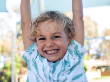 Photo of young boy with curly blond hair hanging from the monkey bars